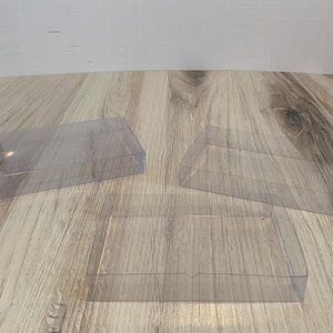 May include: Three clear, rectangular plastic boxes with lids. The boxes are transparent, allowing visibility of the contents. The boxes are empty and sit on a light-colored wooden surface.