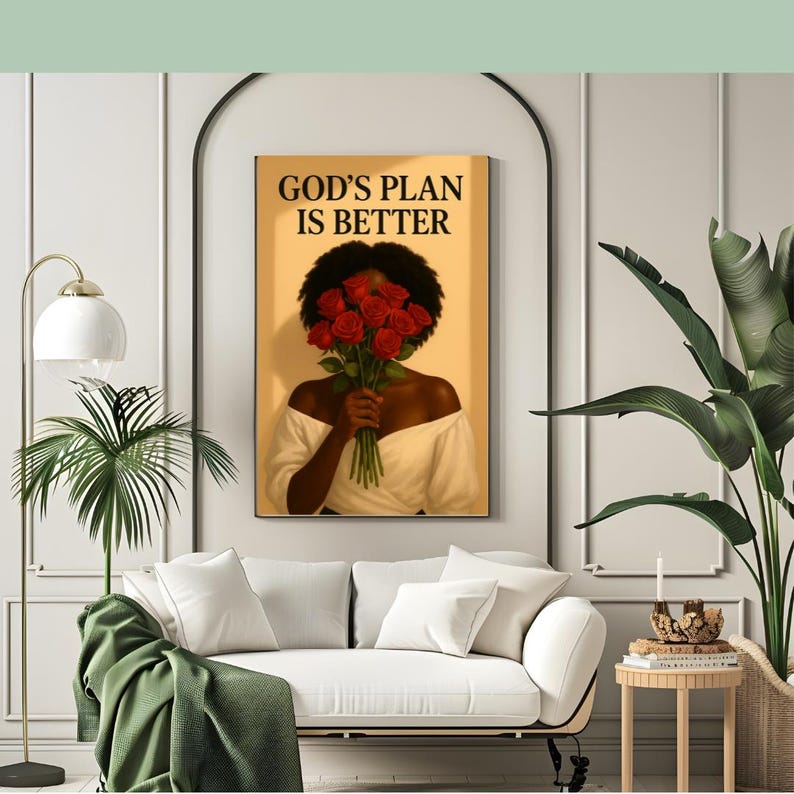 May include: A framed art print featuring a woman holding a bouquet of red roses, with the text "GOD'S PLAN IS BETTER" in black. The woman is wearing a white off-the-shoulder top. The artwork is displayed above a white sofa.