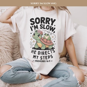 Op de afbeelding: Crèmekleurig T-shirt met een cartoon illustratie van een schildpad en een kikker. De schildpad heeft een bloemenkrans en de kikker zit op zijn rug. Tekst: "SORRY I'M SLOW, HE DIRECTS MY STEPS - PROVERBS 16:9-".