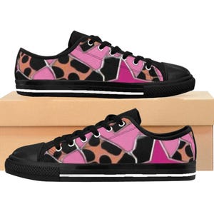May include: Two pairs of low-top canvas trainers. The trainers have a black toe cap and sole with a white stripe. The upper features a pink, black, and orange abstract pattern. The trainers are displayed on a cardboard box.