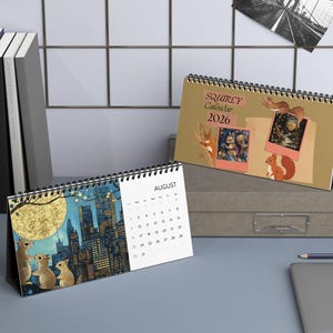 May include: Two desk calendars with spiral binding. One calendar features a cityscape with squirrels, the other displays the text "SQUIRLEY Calendar 2026" and squirrel illustrations. A black and white photo of a bridge is in the background.