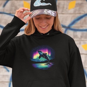 May include: Black hoodie featuring a leaping orca design under the aurora borealis. The hoodie has a front pocket and a drawstring hood. The person is also wearing a black baseball cap with a wave design.