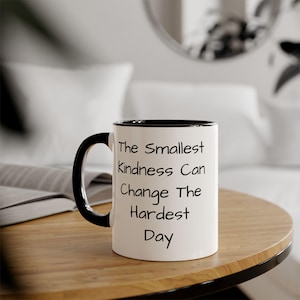 Peut inclure: Mug en céramique blanche avec une anse et un bord noirs. Le mug porte l'inscription "The Smallest Kindness Can Change The Hardest Day" en lettres noires. Le mug est posé sur une table en bois.