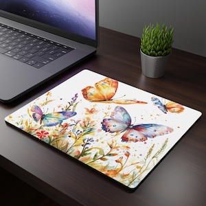 May include: A white mousepad with a watercolor design of butterflies and flowers. The butterflies are in shades of orange, blue, and purple, and the flowers are in shades of yellow, pink, and green.