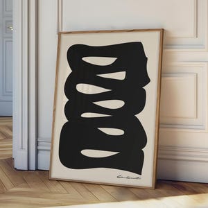 May include: A framed art print featuring a black abstract design on a cream background. The artwork is in a wooden frame and leans against a white wall and door. The print has a minimalist aesthetic, suitable for modern home decor.