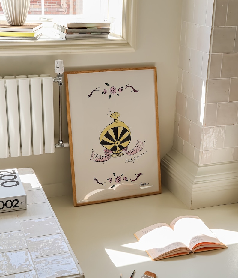 May include: A framed artwork featuring a whimsical illustration of a perfume bottle with a black and yellow design. The artwork includes the word "PARFUM" and decorative floral elements. The frame is made of light-colored wood.