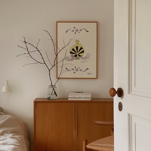 May include: A framed artwork featuring a whimsical perfume bottle design in black, yellow, and pink, with the word "PARFUM" below. The artwork hangs above a wooden cabinet. A glass vase with branches sits on the cabinet.