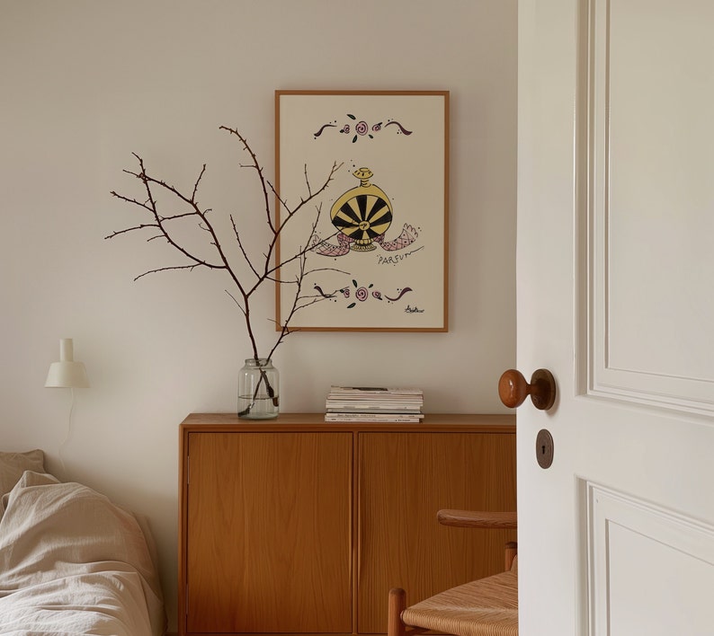 May include: A framed artwork featuring a whimsical perfume bottle design in black, yellow, and pink, with the word "PARFUM" below. The artwork hangs above a wooden cabinet. A glass vase with branches sits on the cabinet.