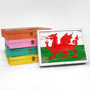 May include: Four wooden boxes stacked on top of each other. The top box has a white background with a red dragon on a green field. The dragon is the Welsh flag. The boxes are painted in different colors: orange, pink, teal, and yellow.