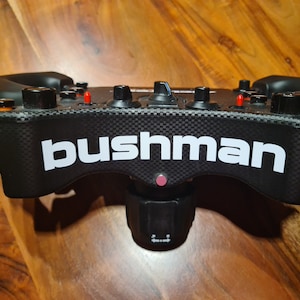 Può includere: Volante da corsa nero con finitura in fibra di carbonio e logo "bushman" bianco. Il volante ha più pulsanti e un pulsante rosso nella parte superiore.