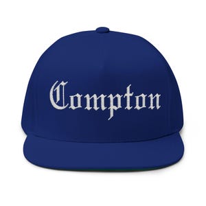 May include: A royal blue baseball cap with a white embroidered inscription that reads "Compton" in a gothic font.