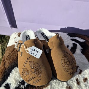 May include: A pair of brown suede clogs with a mushroom design and a white faux fur lining. The clogs have a buckle closure and are size 7.