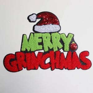 May include: A festive Christmas decoration with the words "MERRY GRINCHMAS" in green and red, topped with a Santa hat. The hat and the text have a textured, sequined appearance, perfect for holiday decor.