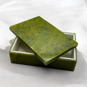 Peut inclure: Une boîte en pierre verte avec un couvercle. La boîte est faite d'une pierre lisse et polie avec un motif vert et blanc naturel. Le couvercle est légèrement entrouvert, révélant l'intérieur blanc de la boîte.