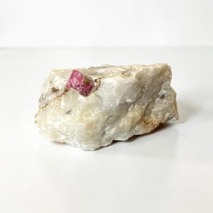 Puede incluir: Un espécimen mineral en bruto y sin pulir que presenta una gran matriz de cuarzo blanco opaco con un único y prominente cristal de color rojo rosado intenso incrustado cerca del borde superior izquierdo, mostrando texturas naturales y formas irregulares.