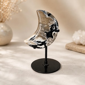 May include: A crescent moon-shaped carving made of polished stone with swirling patterns of beige, cream, and black. The stone is mounted on a thin black metal rod attached to a circular black metal base, creating a vertical display piece.