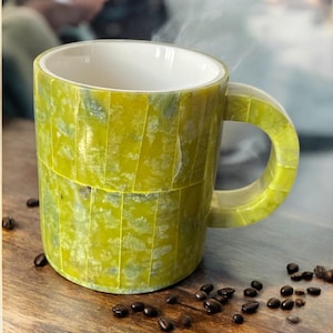 May include: A green mosaic-style coffee mug with a white interior, filled with a steaming beverage. The mug is surrounded by scattered coffee beans on a wooden surface. The mug's handle is also green and has a rectangular shape.