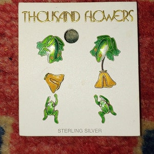 May include: A white card with the text "THOUSAND FLOWERS" in gold lettering. The card features four pairs of sterling silver earrings with green frog and yellow flower designs.