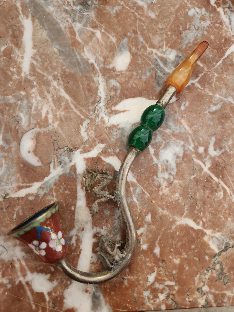 Puede incluir: Una pipa de plata con una cazoleta de esmalte rojo y blanco, cuentas verdes y naranjas y un dise&ntilde;o de drag&oacute;n.