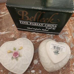 Puede incluir: Una caja de corazón blanca para baratijas con un diseño floral y las palabras "Belleek Fine Parian China Estd. 1857 Hand Crafted in Ireland" en la parte inferior. La caja está hecha de fina porcelana de hueso y tiene un delicado diseño floral.