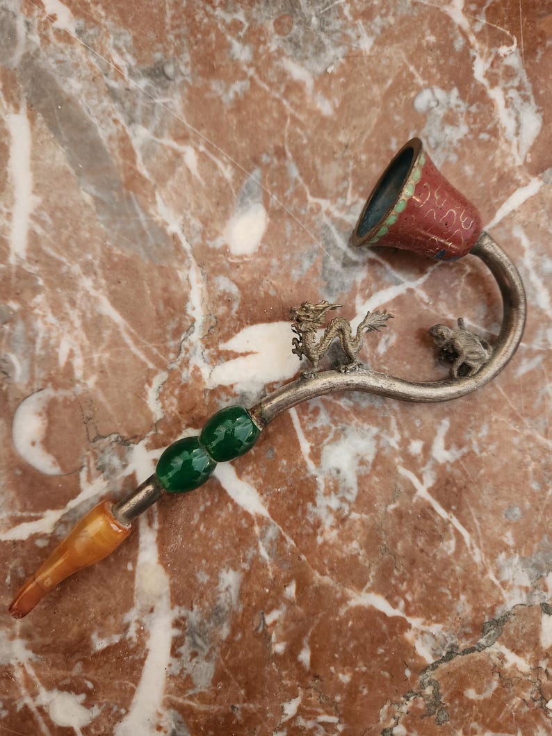Puede incluir: Una pipa de fumar de plata con una figura de drag&oacute;n y una rana. La pipa tiene una cuenta de vidrio verde y una boquilla de &aacute;mbar marr&oacute;n. La cazoleta de la pipa es roja con detalles dorados y el texto "BURMA" escrito en ella.