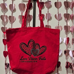 May include: A red tote bag with the words "Love Never Fails" and the Bible verse 1 Corinthians 16:18 printed on it. The bag features three heart designs, two with a leopard print pattern and one with glitter.