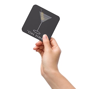 May include: A black square coaster with a white martini glass illustration and the text "tini time" in white.