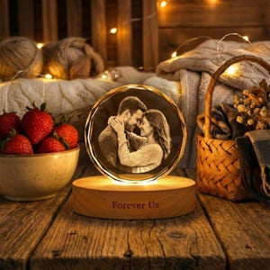 May include: A round, illuminated crystal photo with a couple embracing, set on a wooden base. The base is engraved with "Forever Us." Strawberries in a bowl and a woven basket are in the background, creating a warm, romantic scene.