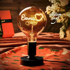 May include: A black metal light bulb base with a clear glass light bulb. The light bulb has a glowing white wire design inside that spells out the name "Emma" with a heart. The text "custom text" is written on the right side of the image.