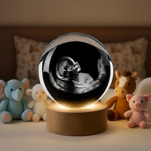K&ouml;nnte beinhalten: Eine klare Kristallkugel mit einem schwarz-wei&szlig;en Ultraschallbild eines F&ouml;tus. Die Kugel steht auf einem Holzsockel, der ein warmes Licht ausstrahlt. Pl&uuml;schtiere, darunter ein Elefant, ein Lamm, ein Pferd und ein Schwein, umgeben den Sockel.