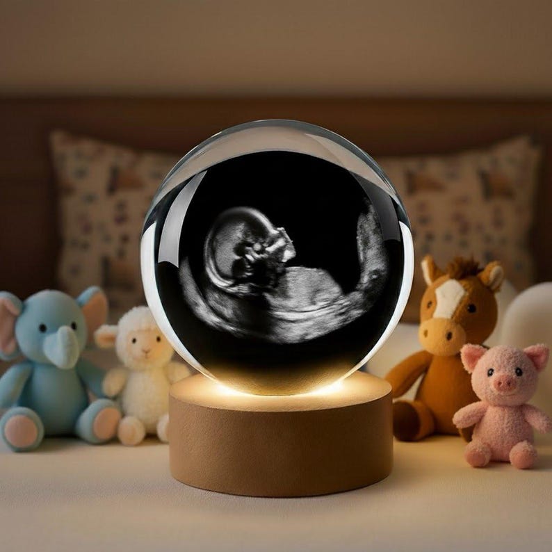 K&ouml;nnte beinhalten: Eine klare Kristallkugel mit einem schwarz-wei&szlig;en Ultraschallbild eines F&ouml;tus. Die Kugel steht auf einem Holzsockel, der ein warmes Licht ausstrahlt. Pl&uuml;schtiere, darunter ein Elefant, ein Lamm, ein Pferd und ein Schwein, umgeben den Sockel.