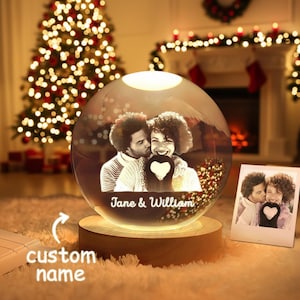 Könnte beinhalten: Eine personalisierte Schneekugel mit einem Foto eines Paares, den Namen "Jane & William". Die Kugel steht auf einem Holzsockel und wird von einem warmen Licht beleuchtet. Ein kleines Foto liegt daneben.