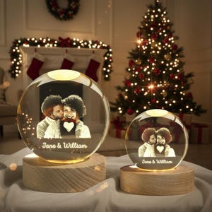 May include: Two illuminated crystal spheres with personalized photos of a couple, set on wooden bases. The spheres have a warm glow, with the names "Jane & William" etched below the image. A Christmas tree and fireplace are in the background.