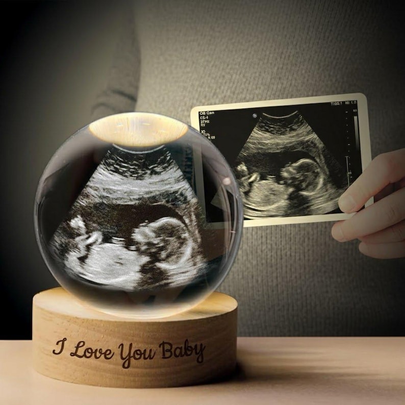 K&ouml;nnte beinhalten: Eine klare Glaskugel mit einem Ultraschallbild im Inneren, die auf einem Holzsockel ruht. Auf dem Sockel steht "I Love You Baby". Ein Ultraschallbild wird daneben gehalten. Ein sentimentales Andenken.