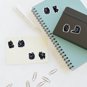 May include: A set of black and white stickers featuring illustrations of Pomeranian dogs. The stickers are on a light blue notebook with a spiral binding and a dark brown notebook.