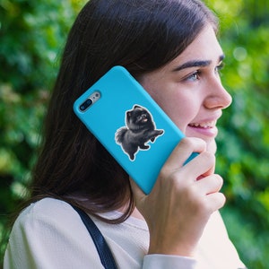 May include: A woman is holding a blue phone case with a black and white dog sticker on it. The dog is a Pomeranian.