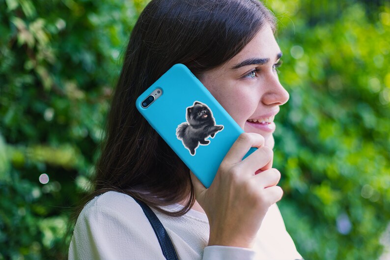 May include: A woman is holding a blue phone case with a black and white dog sticker on it. The dog is a Pomeranian.