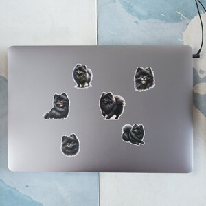 May include: A silver laptop computer with six black and white dog stickers on the lid. The stickers are of a breed of dog that looks like a Pomeranian.