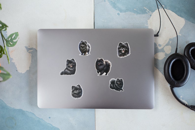 May include: A silver laptop computer with six black and white dog stickers on the lid. The stickers are of a breed of dog that looks like a Pomeranian.