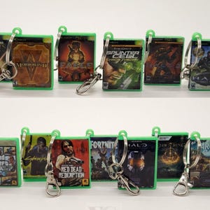 May include: A set of 10 green and black video game keychains featuring popular game titles like Halo, Red Dead Redemption, and Grand Theft Auto. Each keychain is a replica of a video game case with a silver metal ring.