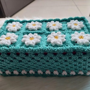 May include: A teal crocheted tissue box cover with white daisy flowers. The cover has a crocheted border with white accents.