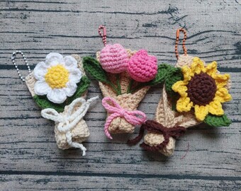 Può includere: Tre bouquet di fiori all'uncinetto. Uno presenta una margherita bianca con centro giallo, un altro tulipani rosa e l'ultimo un girasole giallo. Ogni bouquet è avvolto in filo beige e legato con un nastro. Hanno una piccola catena attaccata.