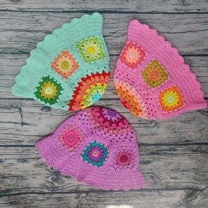 Puede incluir: Tres sombreros de pescador tejidos a mano con motivos de cuadrados de la abuela coloridos. Un sombrero es verde menta claro, otro es rosa brillante y otro es morado lavanda oscuro. Cada sombrero muestra patrones florales en varios colores.