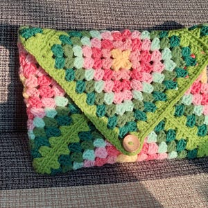 Może przedstawiać: Zielona, różowa i biała poduszka wykonana na szydełku z guzikiem w środku. Poduszka wykonana jest w wzorze granny square.