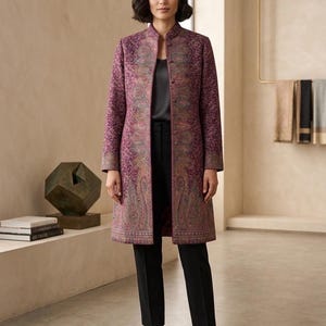 May include: A long, patterned coat in shades of purple, pink, and brown. The coat features a mandarin collar and a detailed paisley design. The model is wearing black pants and brown flats. The coat is a statement piece.