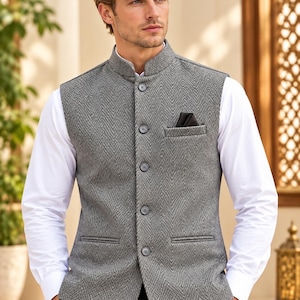 May include: A grey herringbone patterned vest with a mandarin collar, five front buttons, and a chest pocket containing a dark pocket square, worn over a crisp white long-sleeved button-down shirt and black trousers.