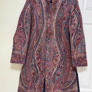 May include: A long-sleeved, knee-length coat featuring a mandarin collar and a front button closure. The garment is decorated with an intricate, multi-colored paisley pattern in shades of deep red, burgundy, navy blue, and teal on a textured fabric.