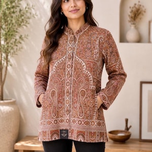 May include: A long-sleeved jacket in a warm brown tone featuring an intricate paisley pattern in shades of cream, rust, and dark brown. The jacket has a mandarin collar, a central button-down placket, and two side pockets, paired with black pants.