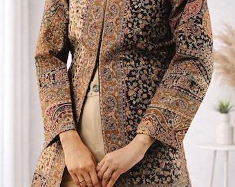 May include: A long-sleeved jacket featuring a high collar and an intricate, multi-colored paisley and floral pattern in shades of black, gold, rust, and cream. The garment has a straight-cut silhouette and is worn over plain beige trousers.
