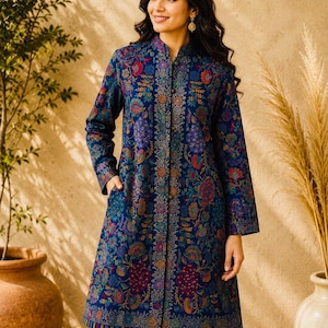 May include: A long-sleeved, knee-length tunic in a deep navy blue fabric featuring a dense, multicoloured floral pattern in shades of red, orange, purple, and gold. The garment has a mandarin collar, a central button placket, and side pockets.